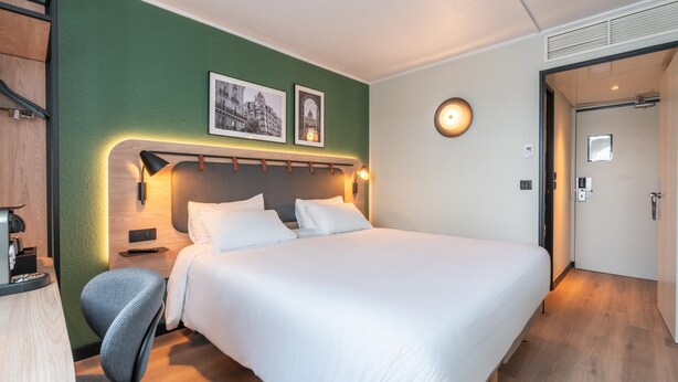 A hotel room with a large bed, a desk with a chair, a wall-mounted lamp, and two framed pictures. - Campanile PRIME - Paris Gennevilliers Barbanniers (Gennevilliers)