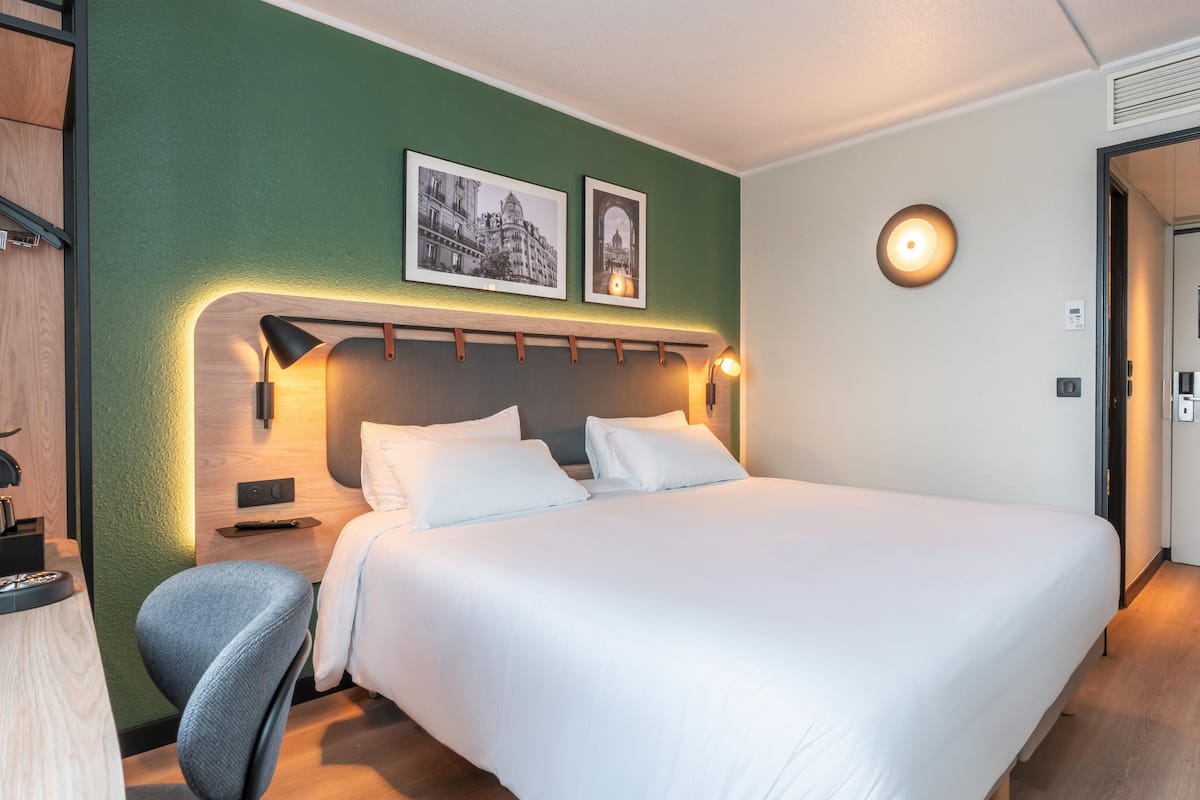 Une chambre d’hôtel avec un grand lit, un bureau avec une chaise, une lampe fixée au mur et deux tableaux encadrés.