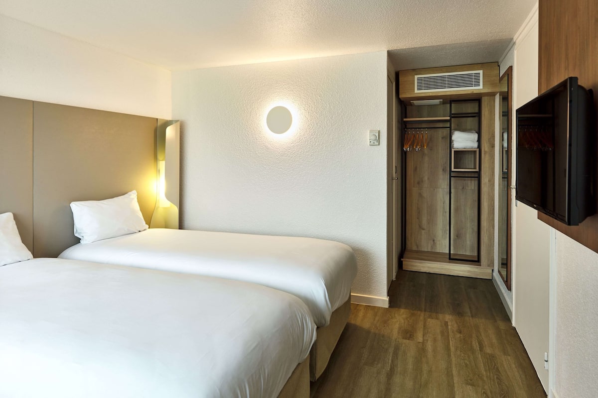 A hotel room with two beds, a television, a wardrobe, and a circular light fixture on the wall.