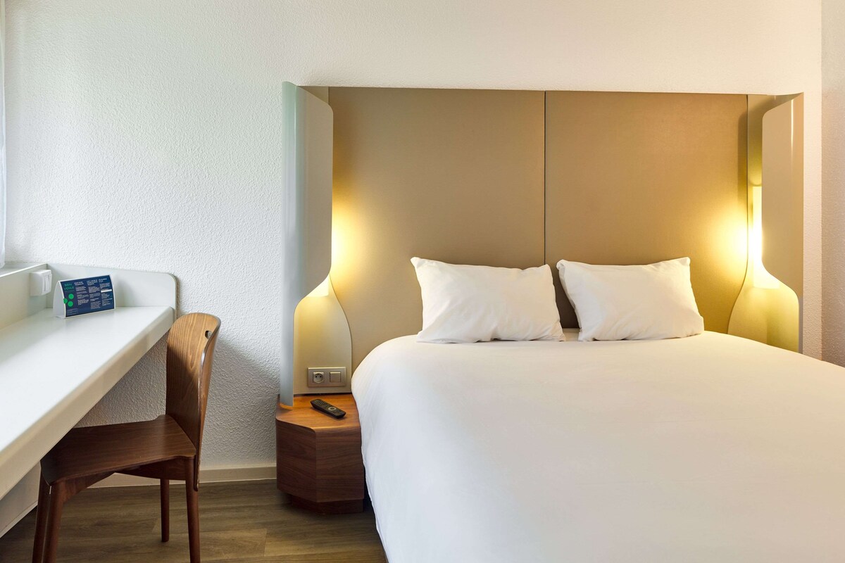 A hotel room with a bed, bedside table, chair, and a control panel.