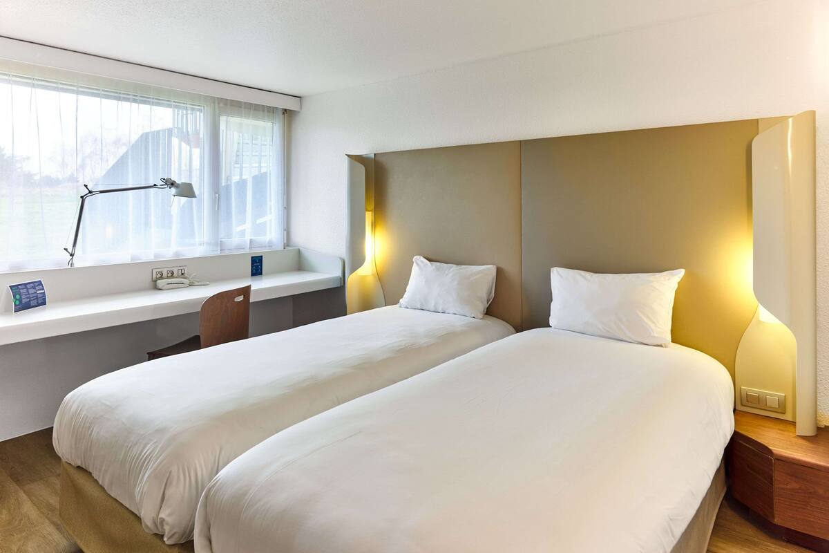 A hotel room with two beds, a desk with a lamp, and a window with curtains.