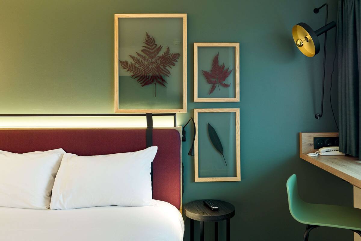 A hotel room with a bed, a desk with a chair, and framed artwork on the wall.