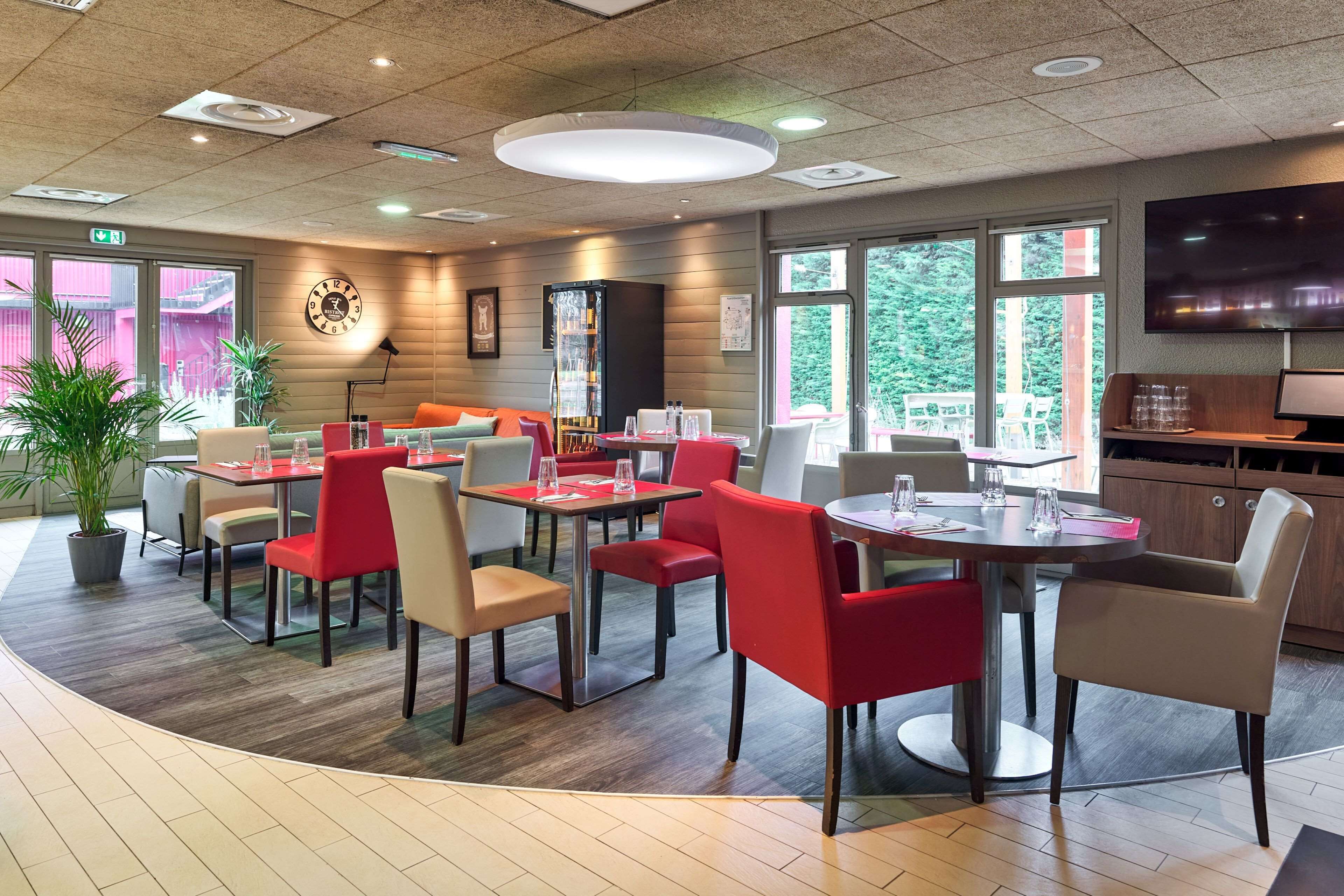 Un espace repas moderne avec des tables et des chaises rondes, une horloge au mur et de grandes fenêtres donnant sur un paysage verdoyant.