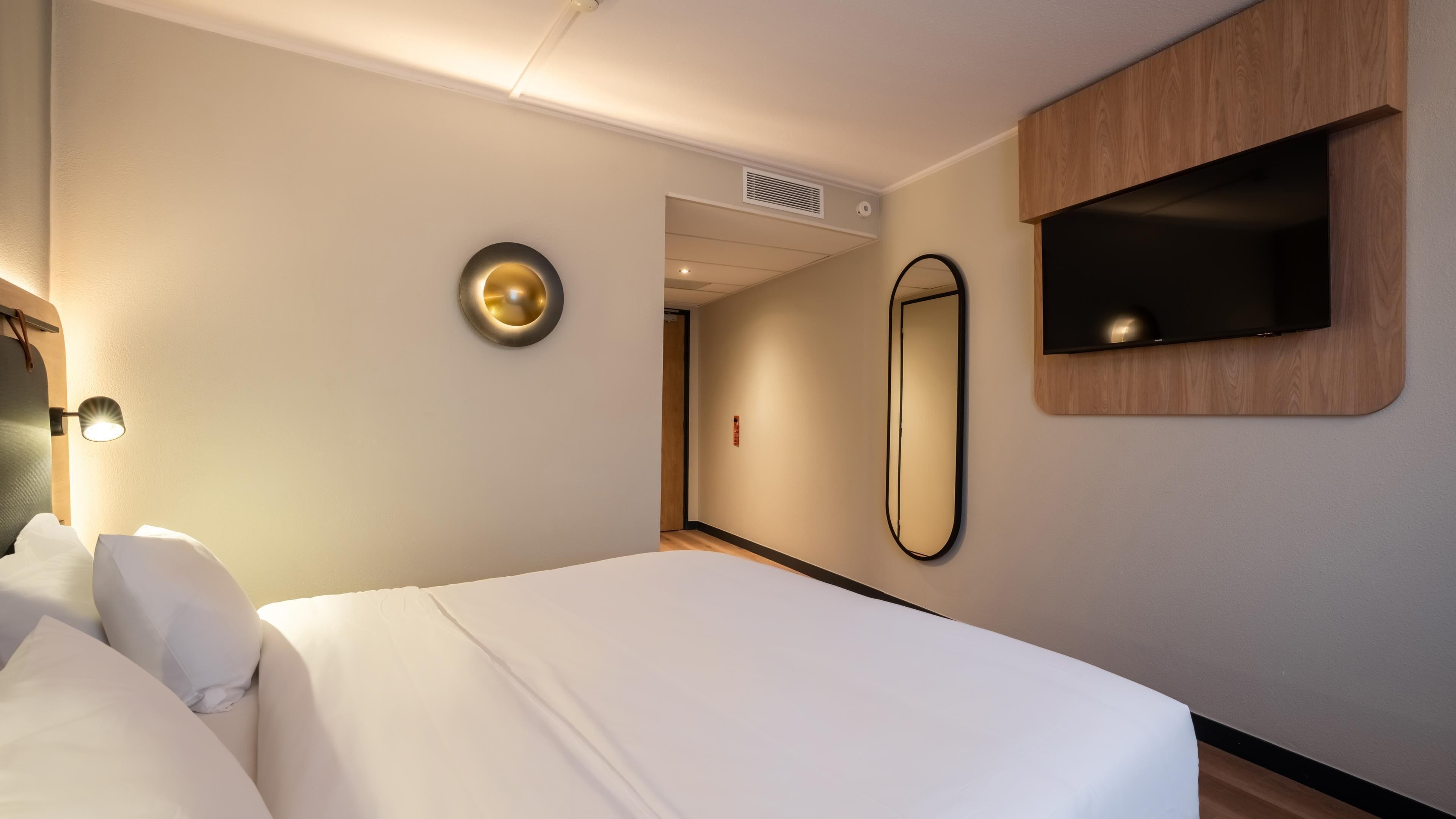 A hotel room with a bed, a wall-mounted mirror, a TV, and a wall-mounted light fixture.