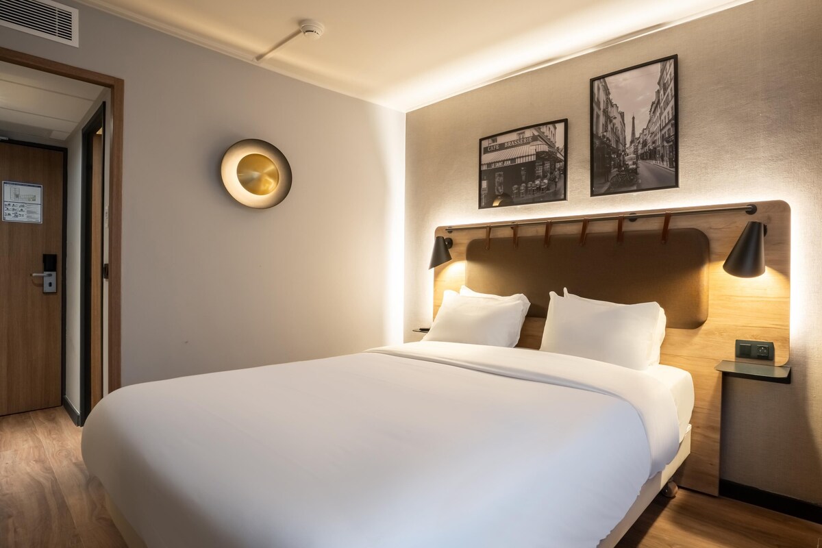 A hotel room with a large bed, a headboard with a light fixture, a wall-mounted clock, and two framed pictures.
