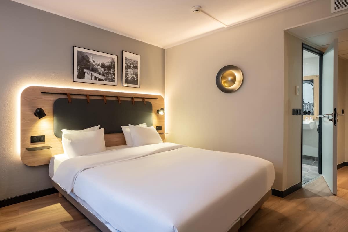 A modern hotel room with a large bed, a headboard with built-in lighting, two framed pictures on the wall, and a round wall-mounted light fixture.