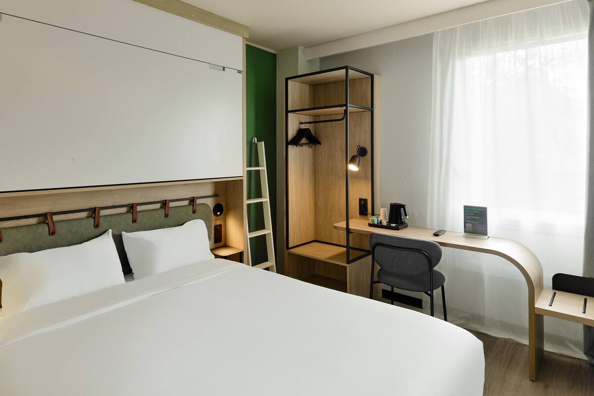 A hotel room with a bed, a desk, a chair, a ladder, and a small shelf with a coffee maker and a sign.