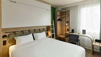 Una camera d'albergo con un letto, una scrivania, una sedia, una scala e un piccolo ripiano con una macchina del caffè e un cartello.