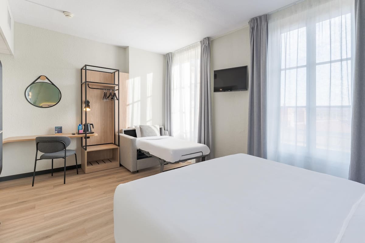A hotel room with a bed, a desk, a chair, a TV, and large windows with curtains.