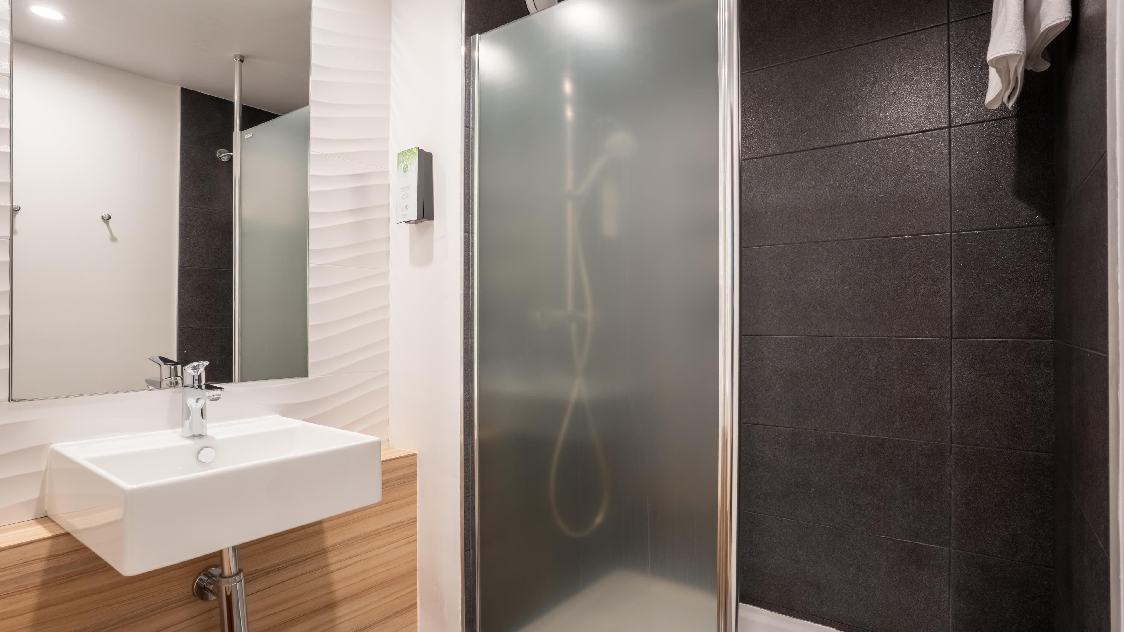 A modern bathroom with a white sink, a glass shower enclosure, and dark tiled walls.