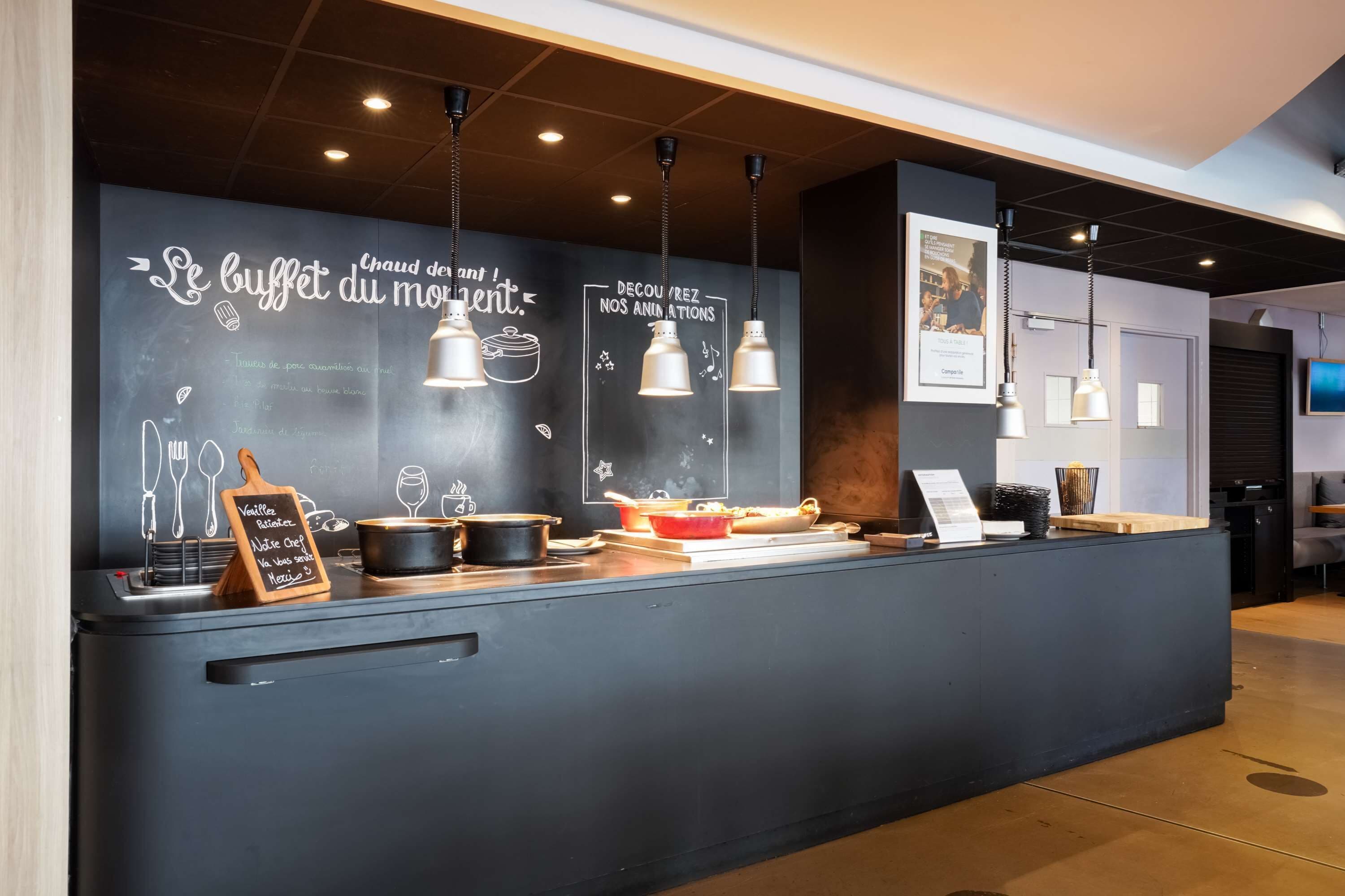 Una encimera de cocina moderna con un menú en una pizarra, luces colgantes y una pared con pizarra que muestra artículos y dibujos del menú.