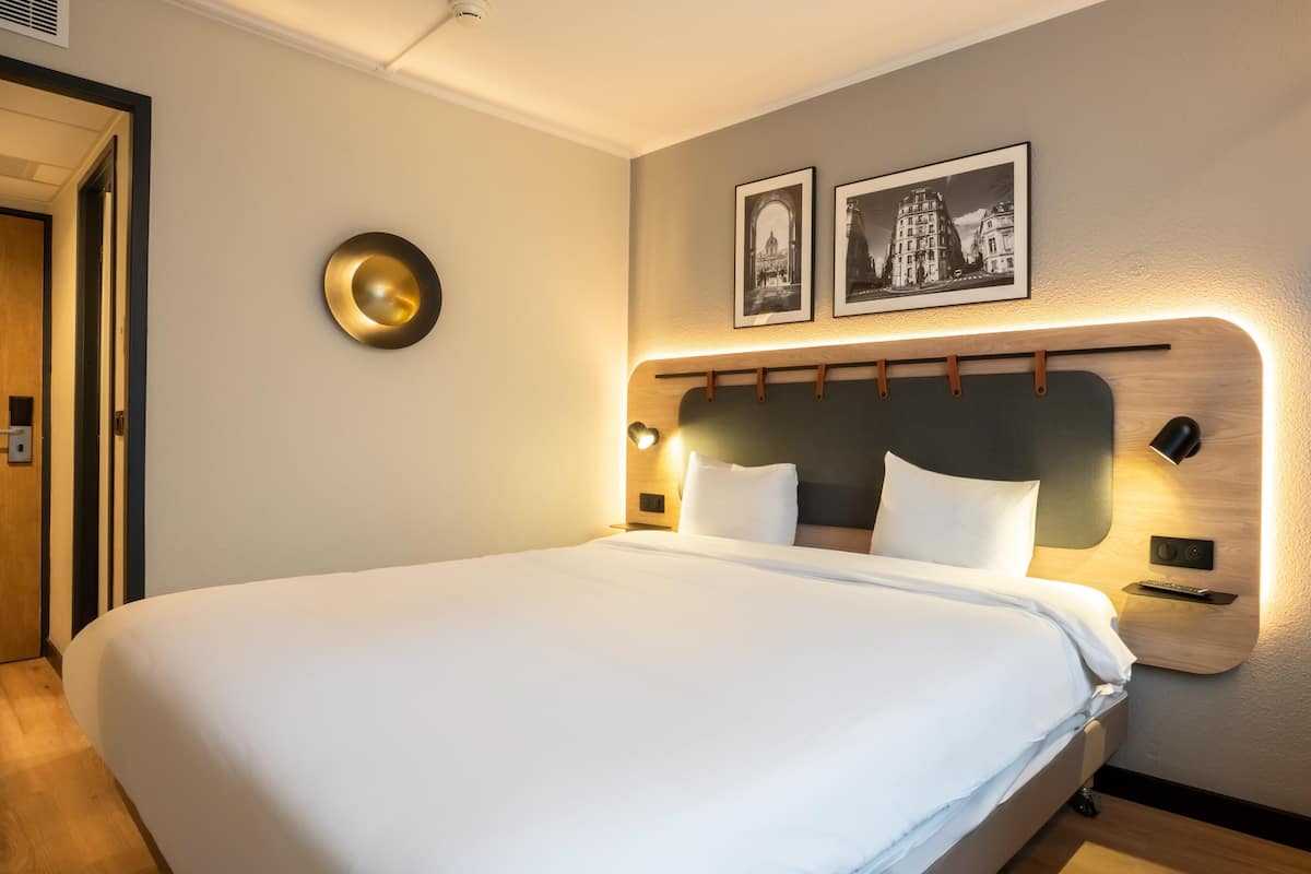 A hotel room with a large bed, a headboard with integrated lighting, a desk with a chair, and two framed pictures on the wall.
