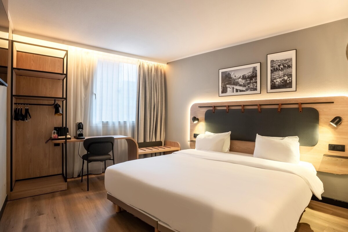 A hotel room with a large bed, a desk with a chair, a wardrobe, and two framed pictures on the wall.
