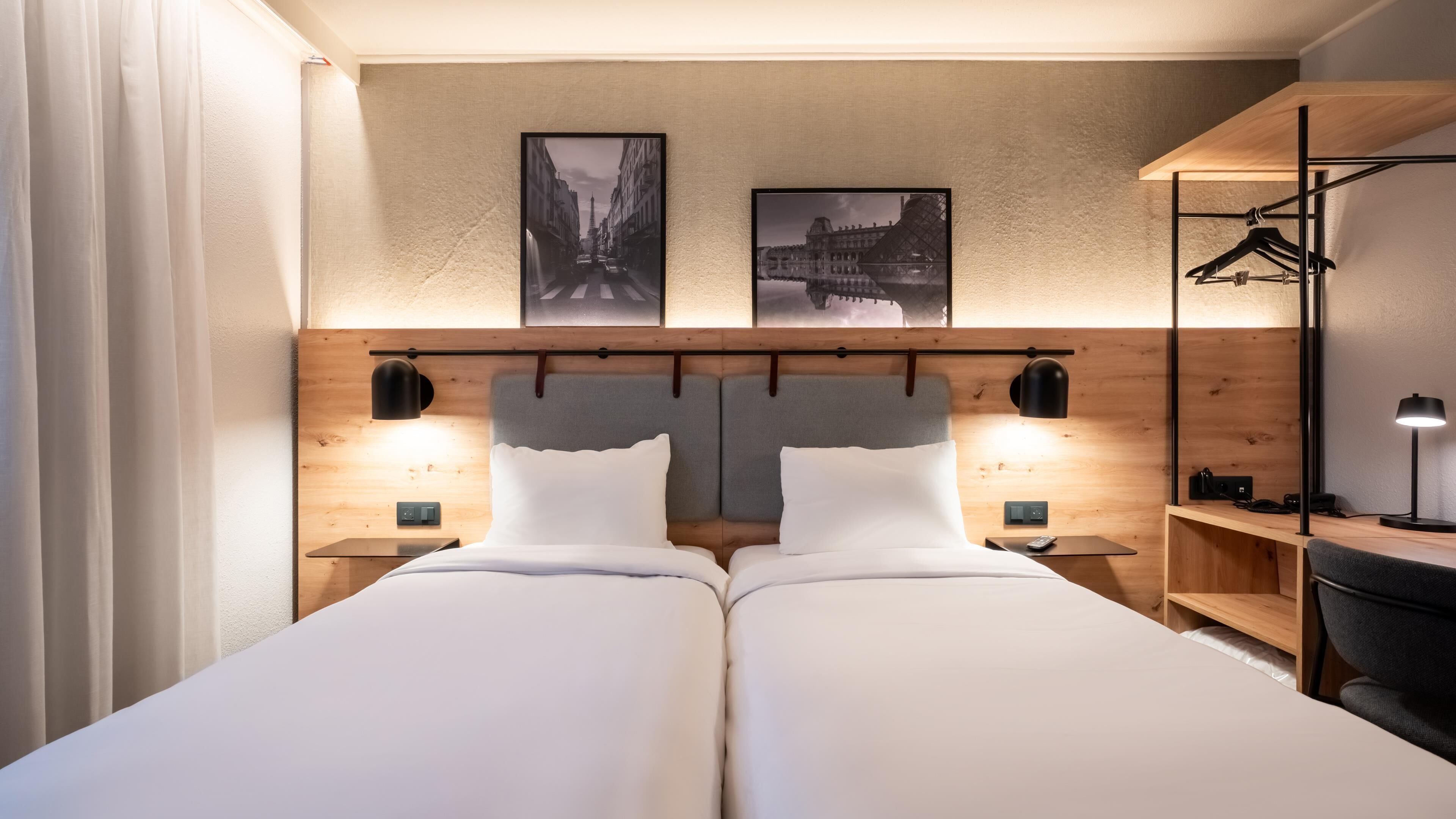 Habitación de hotel con dos camas, cabecero de madera, dos lámparas de pared y dos cuadros enmarcados sobre las camas.