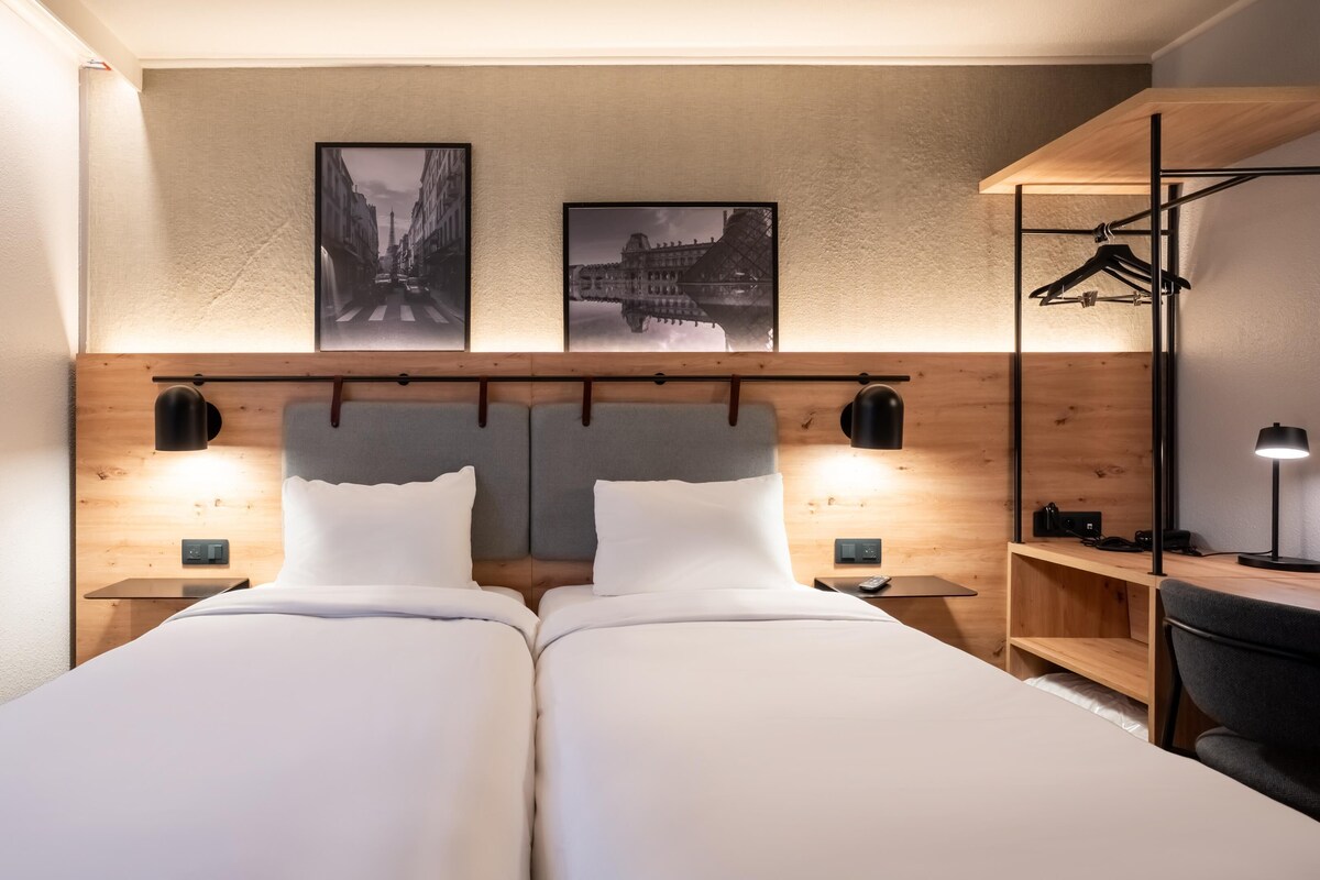 A hotel room with two beds, a wooden headboard, two wall-mounted lamps, and two framed pictures above the beds.