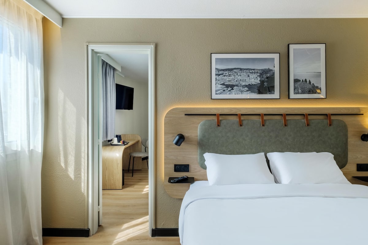 A hotel room with a bed, a desk, a chair, a TV, and two framed pictures on the wall.