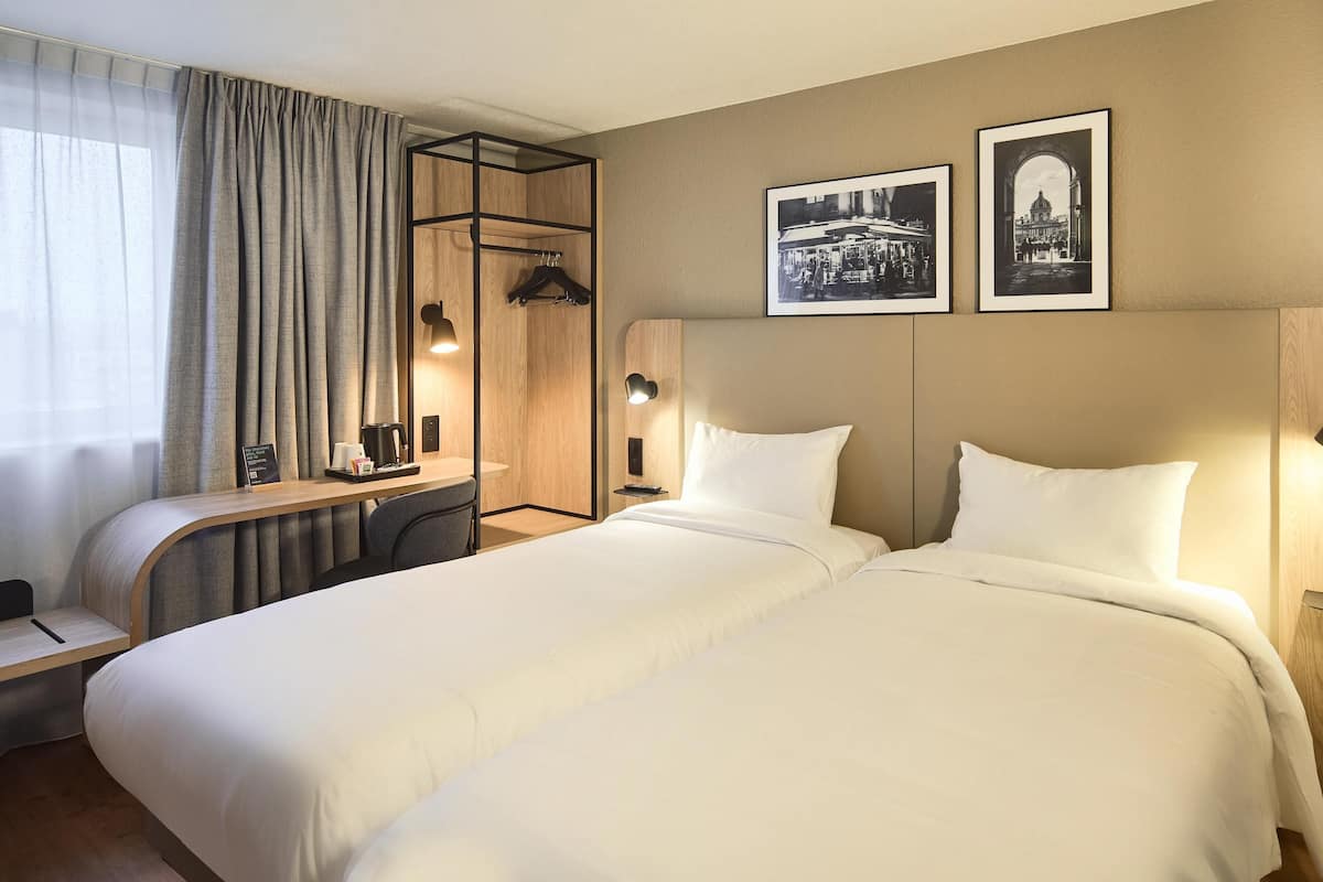 A hotel room with a bed, a desk, a chair, a coffee maker, and two framed pictures on the wall.