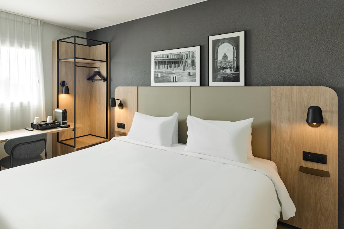 A hotel room with a large bed, a desk with a chair, a coat rack, and two wall-mounted lamps.