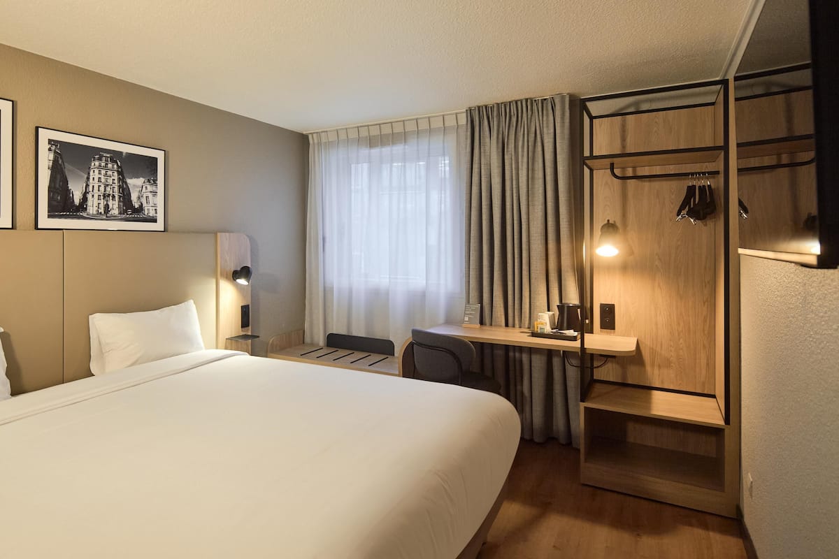 A hotel room with a bed, a desk, a chair, a coat rack, and two framed pictures on the wall.
