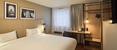 Una habitación de hotel con una cama grande, un escritorio con silla, un perchero y dos cuadros en la pared.