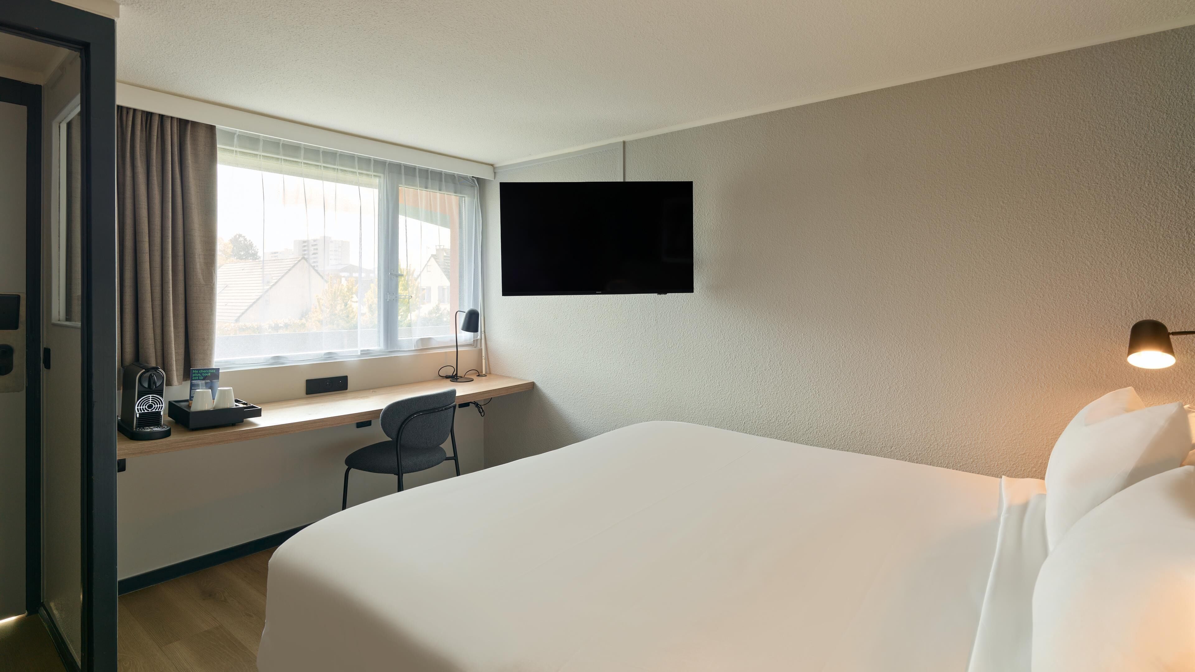 A hotel room with a bed, a desk with a chair, a TV on the wall, and a window with curtains.