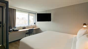 A hotel room with a bed, a desk with a chair, a TV on the wall, and a window with curtains.