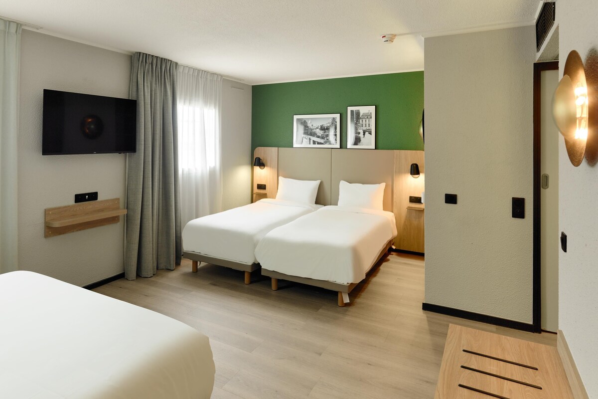 Habitación de hotel con dos camas, un televisor, una estantería de madera y una pared de acento verde.