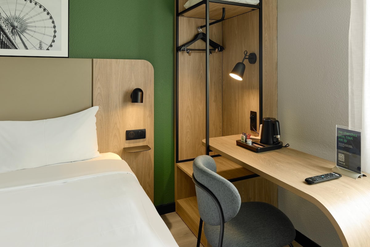 A hotel room with a bed, a desk with a chair, a coffee maker, a hanging light, and a framed picture on the wall.