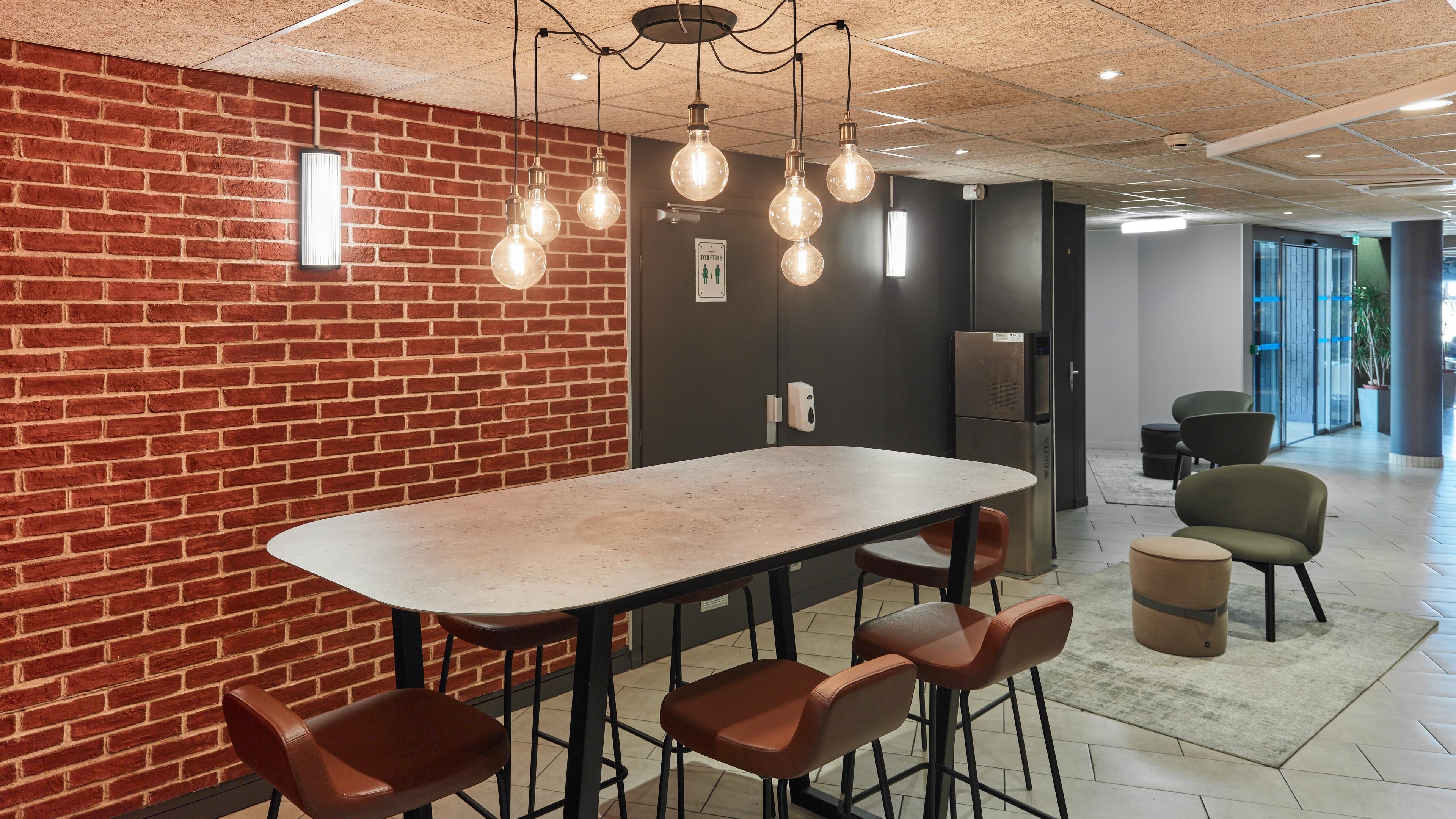 Interior moderno com parede de tijolos, uma mesa comprida com cadeiras, lâmpadas penduradas, uma geladeira e uma placa indicando um banheiro.