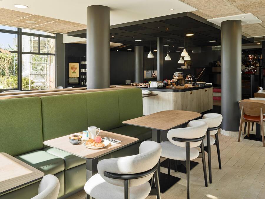 A modern cafe with green booths, wooden tables, and white chairs. There is a coffee machine and a counter with various items.