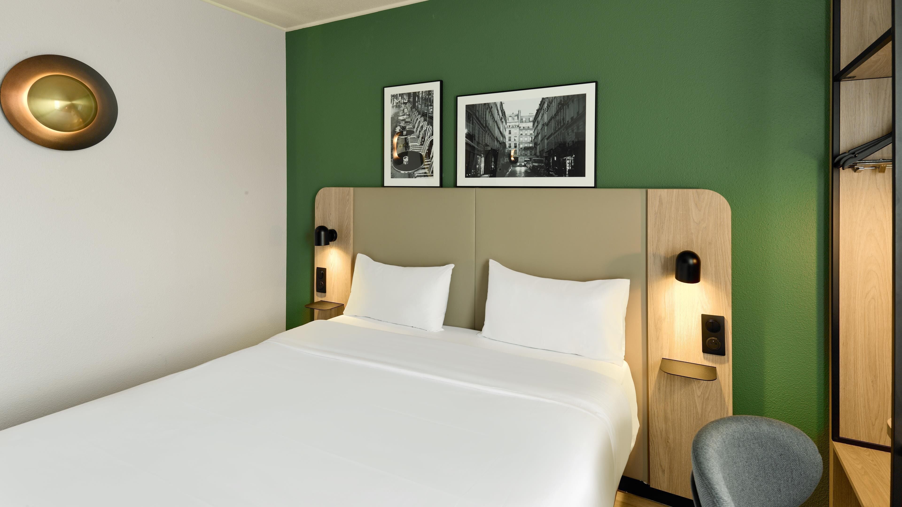 A hotel room with a bed, two wall-mounted picture frames, a round wall light, and a wooden headboard.