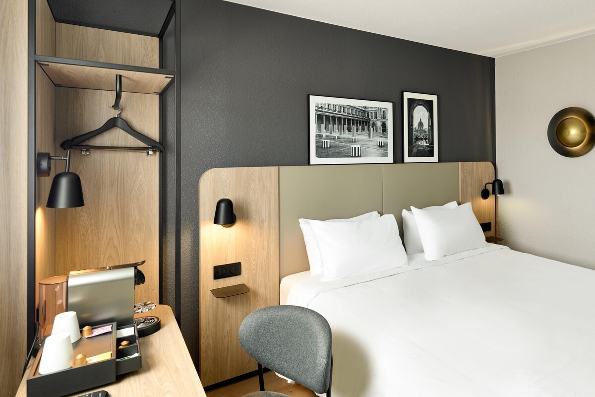 Habitación de hotel con cama, mesita de noche, silla, escritorio, televisión y lámparas de pared.