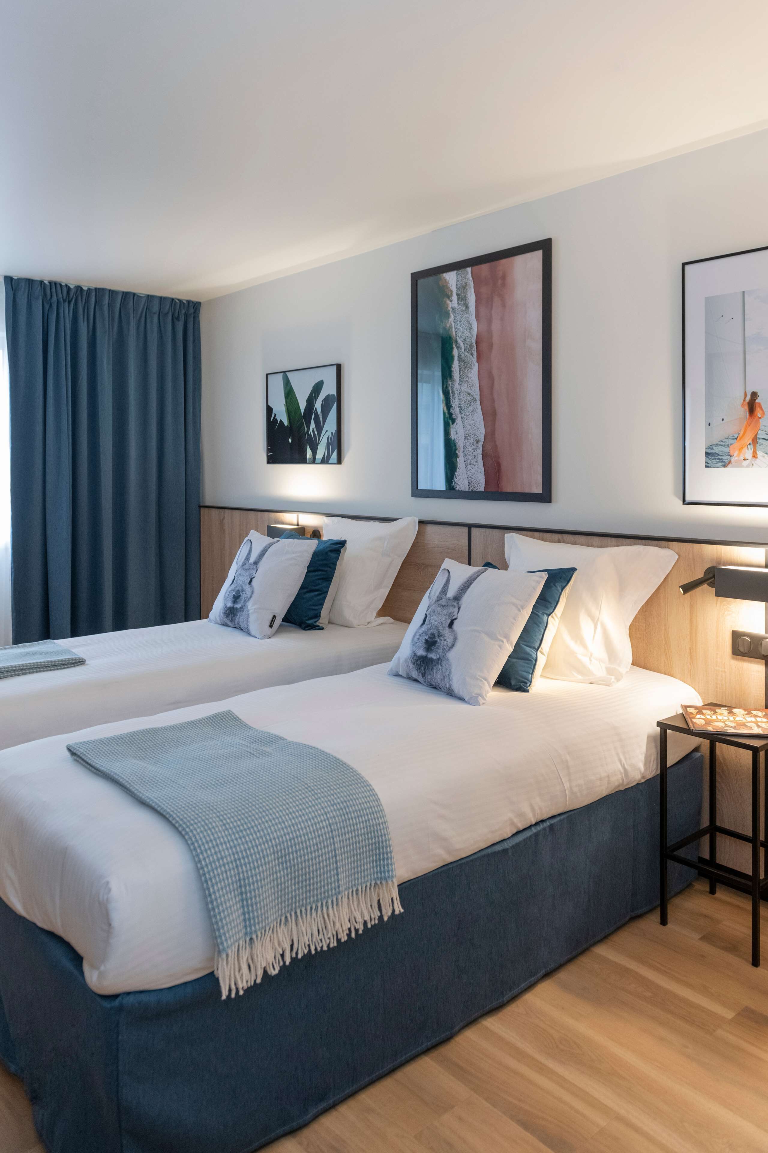 A hotel room with two beds, a small table, and artwork on the walls.