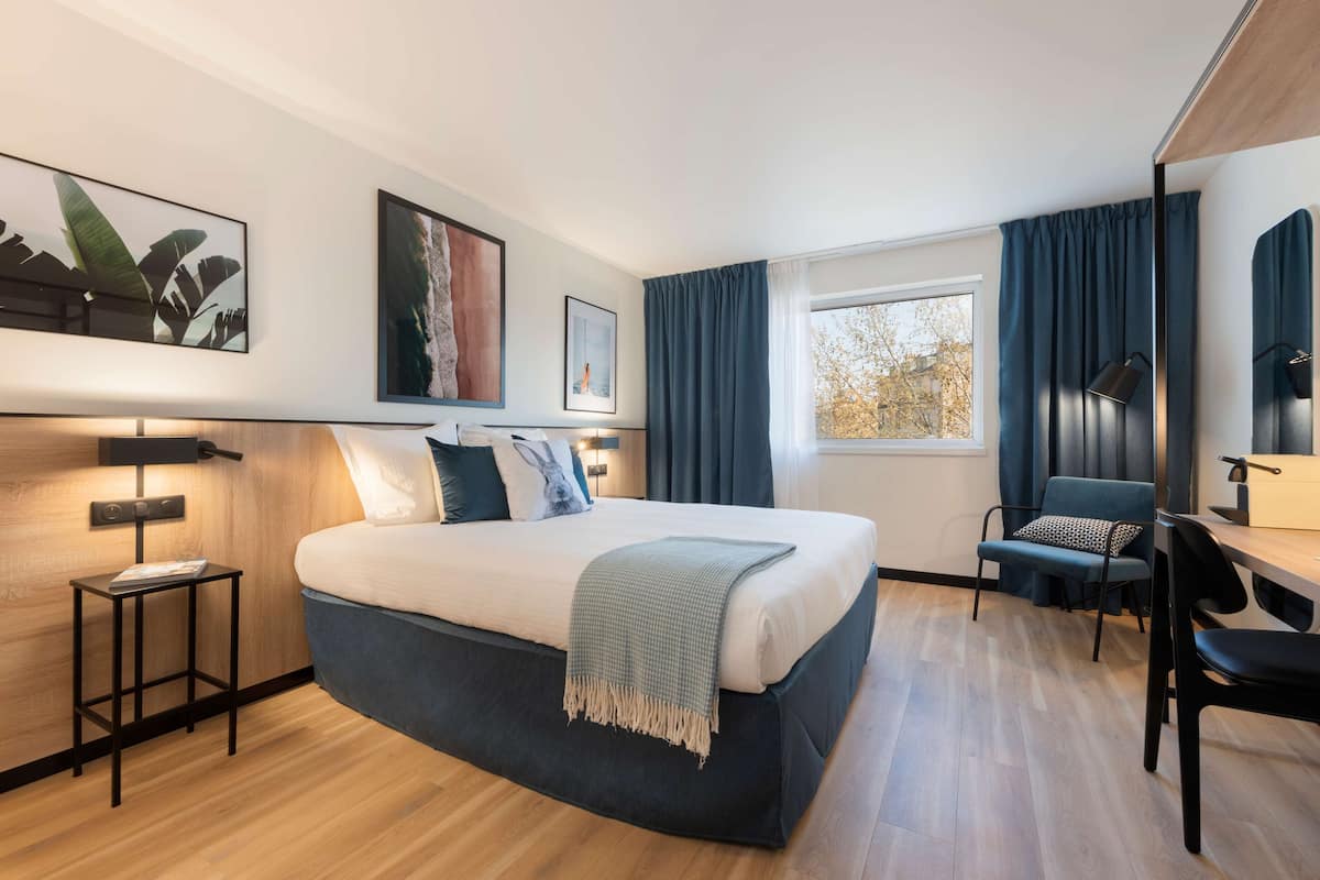 A modern hotel room with a large bed, a desk, and a chair. There are two side tables, a window with curtains, and artwork on the walls.