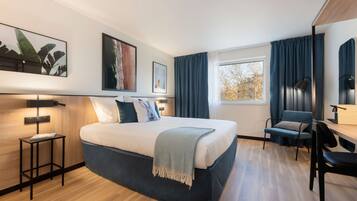 Une chambre d’hôtel moderne avec un grand lit, un bureau et une chaise. On y trouve deux tables de chevet, une fenêtre avec des rideaux et des œuvres d’art accrochées aux murs.