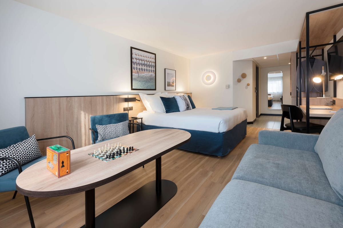 A modern hotel room with a bed, a sofa, a desk, and a chessboard.