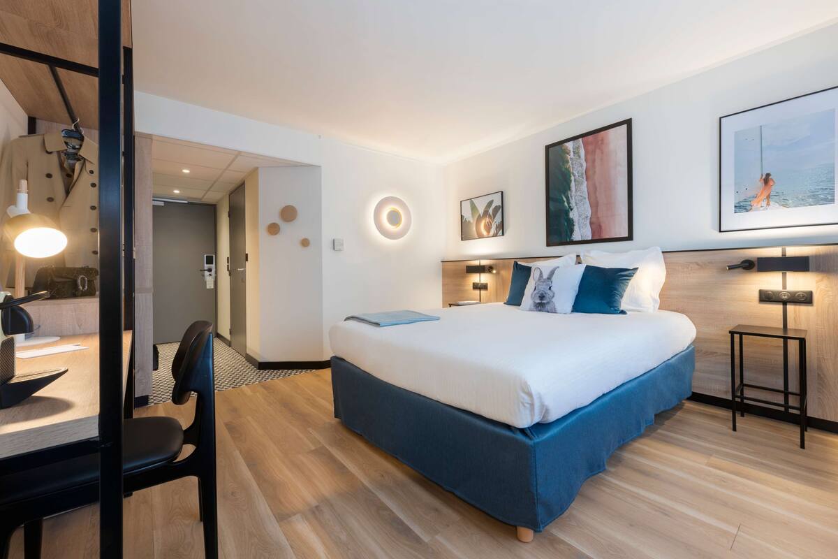 A modern hotel room with a large bed, a desk, and a chair. There are wall-mounted lights and artwork.