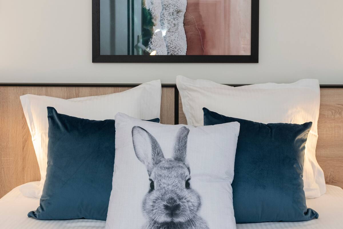 A bed with white bedding and two pillows, one with a rabbit design. Above the bed, there is a framed picture of a coastal scene.