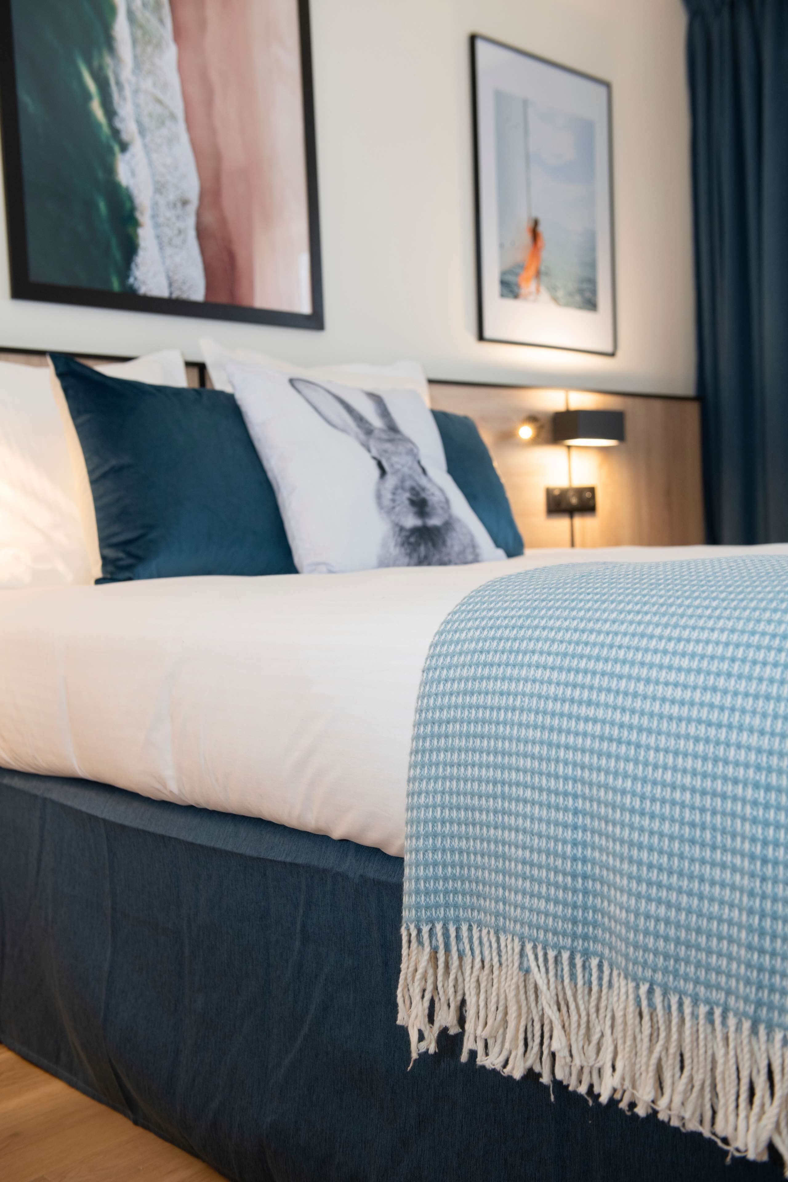 Een net opgemaakt bed met een blauw-wit geruit dekbed, een kussen met een konijntjesprint en een lijst met een foto van een strand.