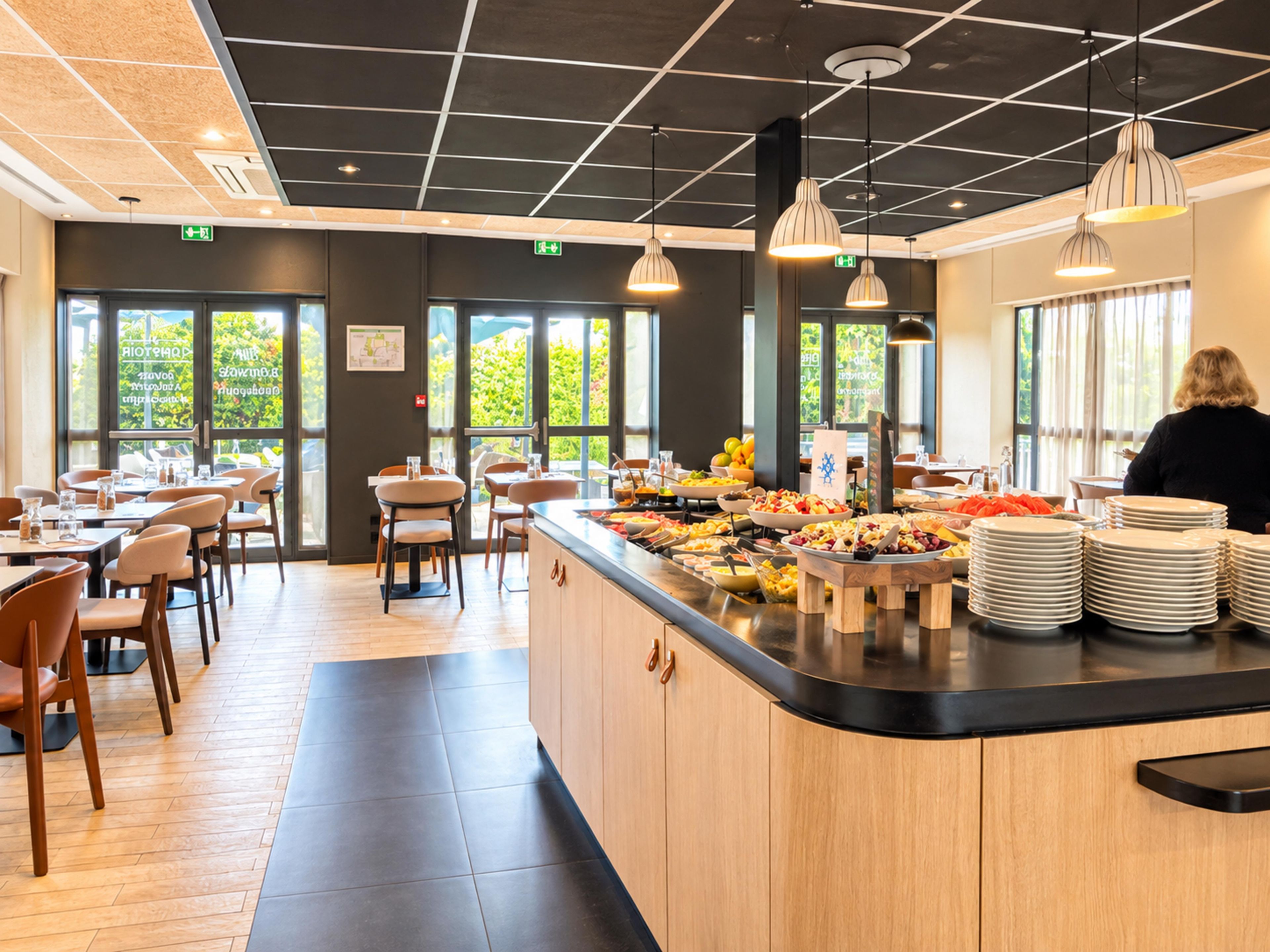 Un espace buffet comprenant plusieurs présentoirs de nourriture, des tables avec des chaises et un comptoir où sont empilées les assiettes.