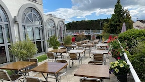 An outdoor patio with tables and chairs, a building with arched windows, and a garden with various plants. - Campanile PRIME - Villejuif (Villejuif)