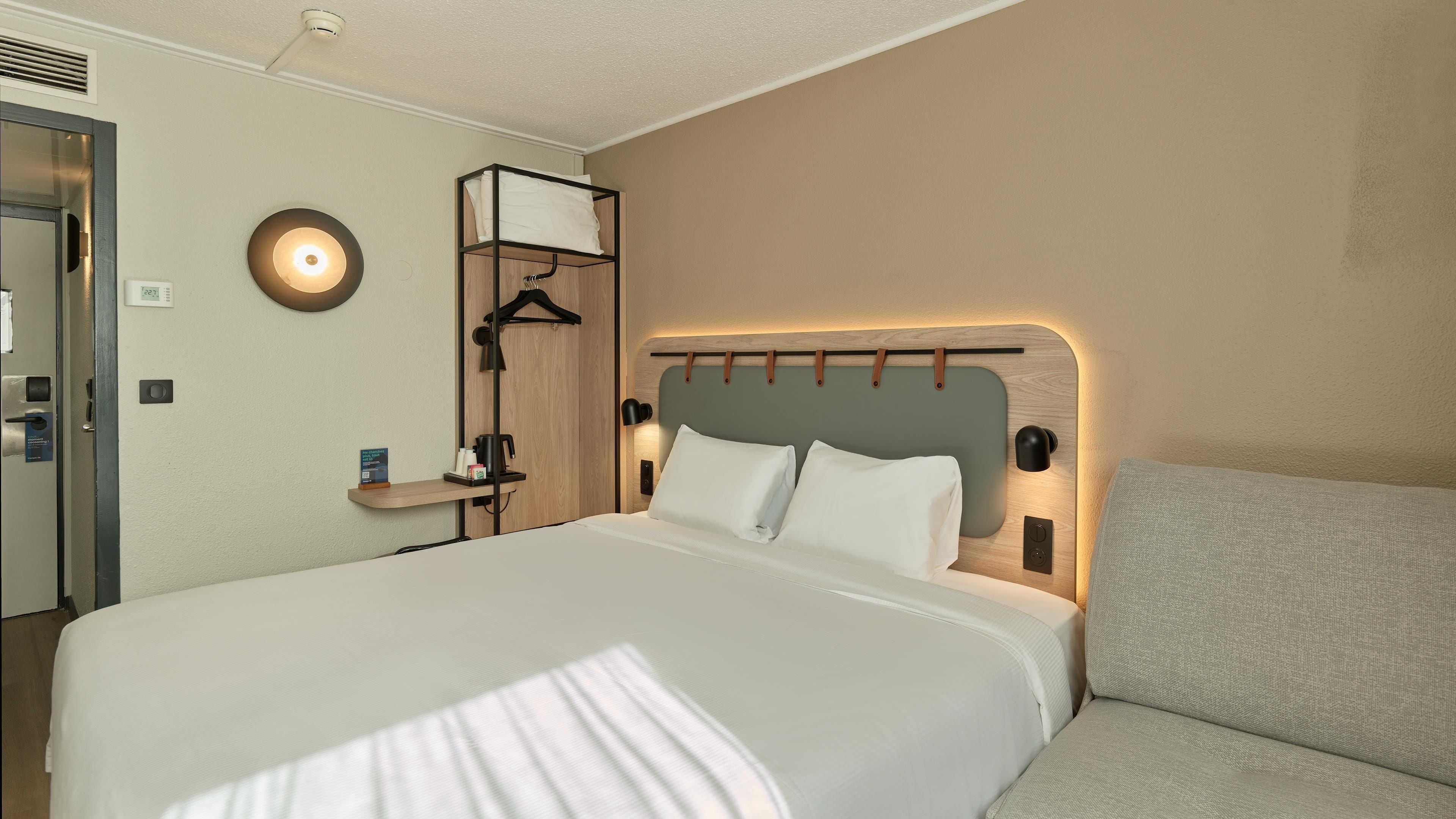 A hotel room with a bed, bedside table, lamp, and a wall-mounted heater.
