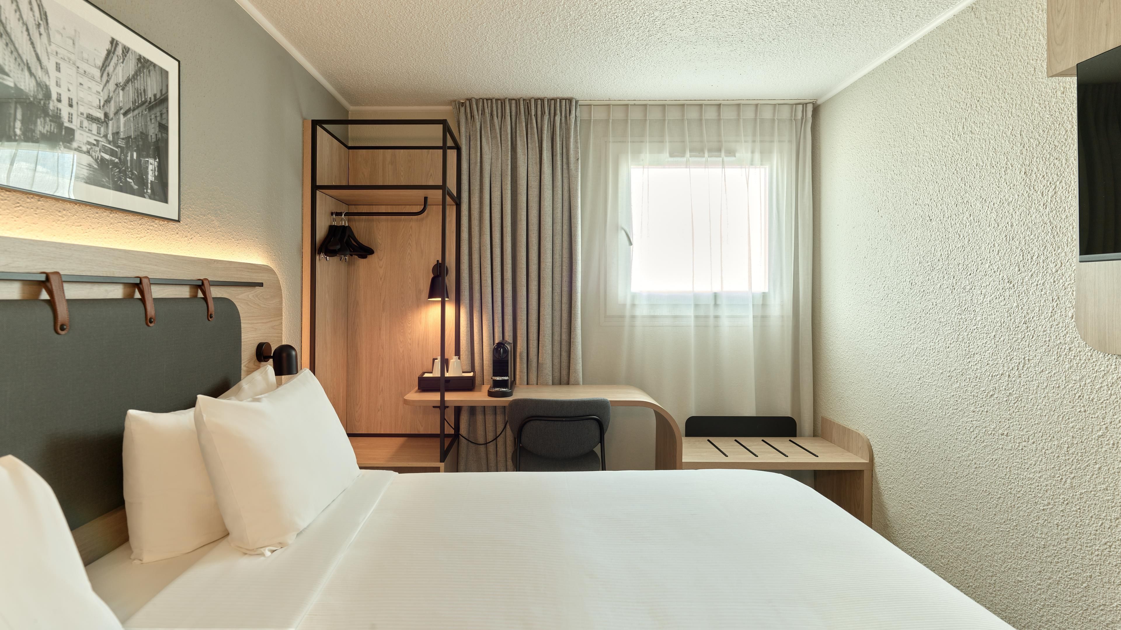 Une chambre d’hôtel comprenant un lit, un bureau, une chaise, un porte-manteau, une lampe et une fenêtre avec des rideaux.