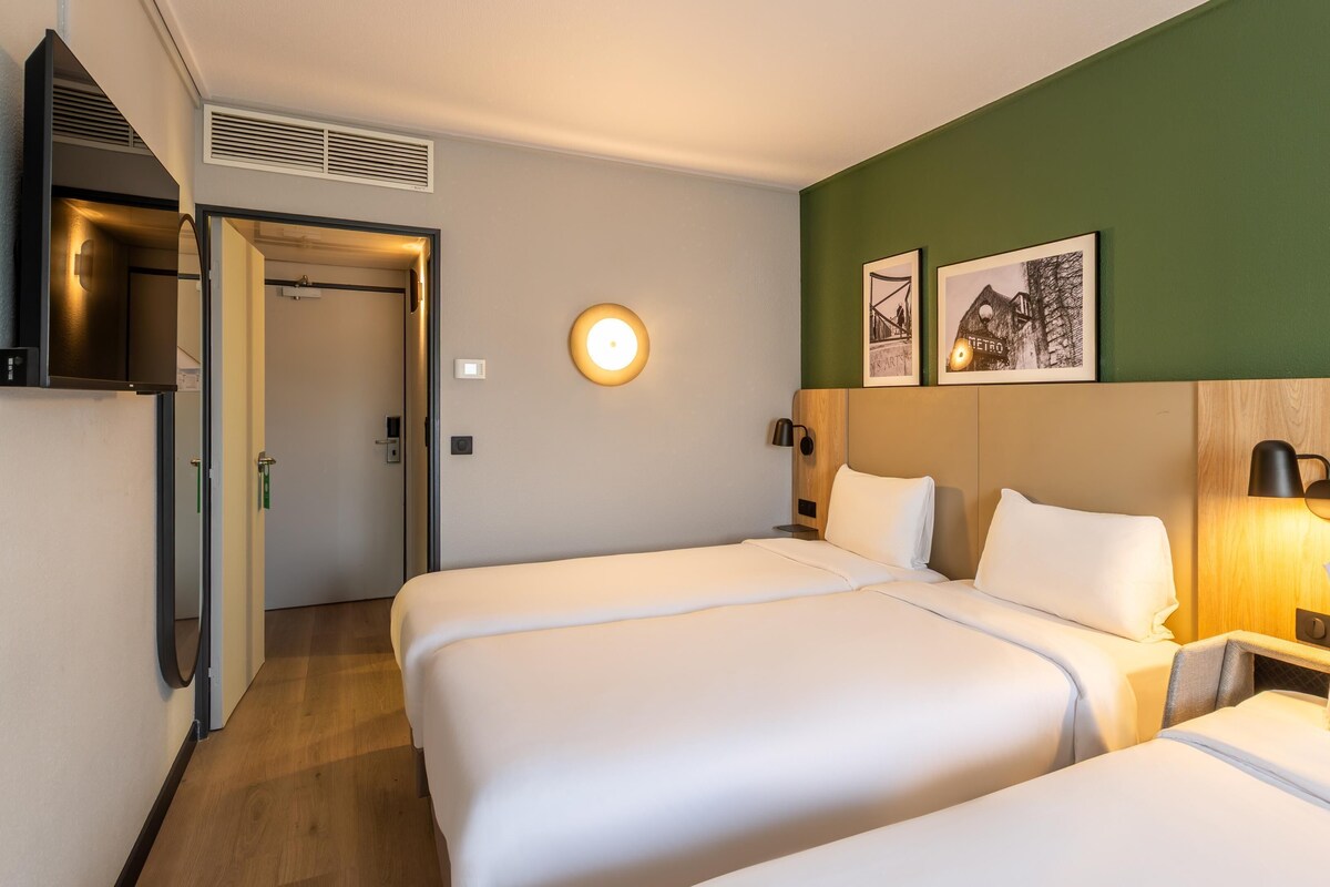 A hotel room with two beds, a door, a wall-mounted light, and two framed pictures.