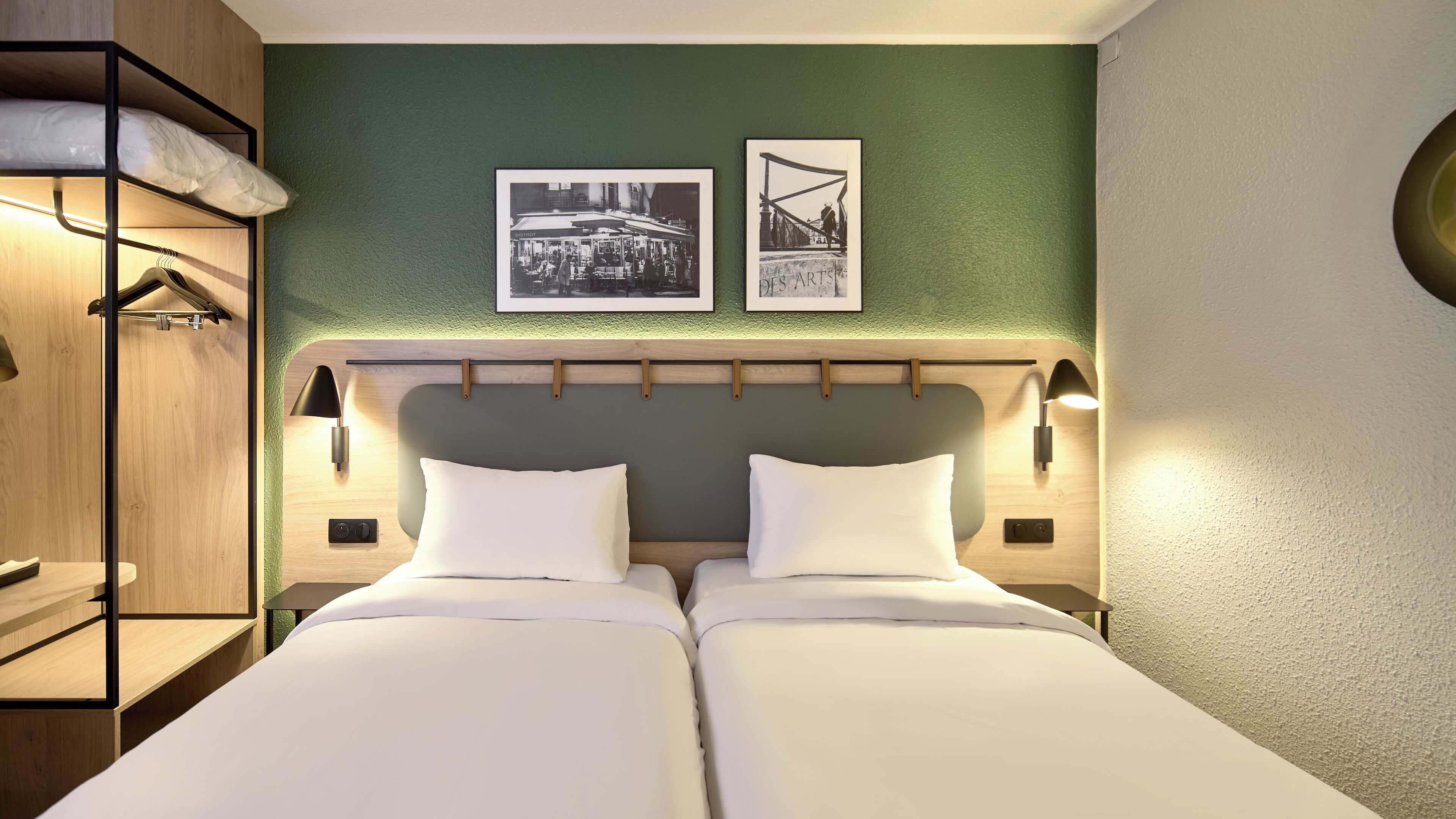 A hotel room with two beds, a wooden wardrobe, and two framed pictures on the wall.