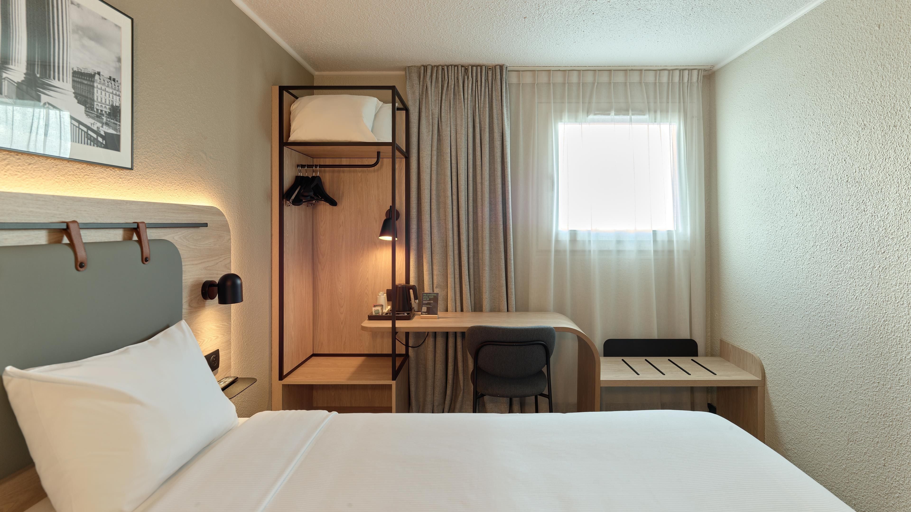 A hotel room with a bed, a desk with a chair, a wardrobe, and a window with curtains.