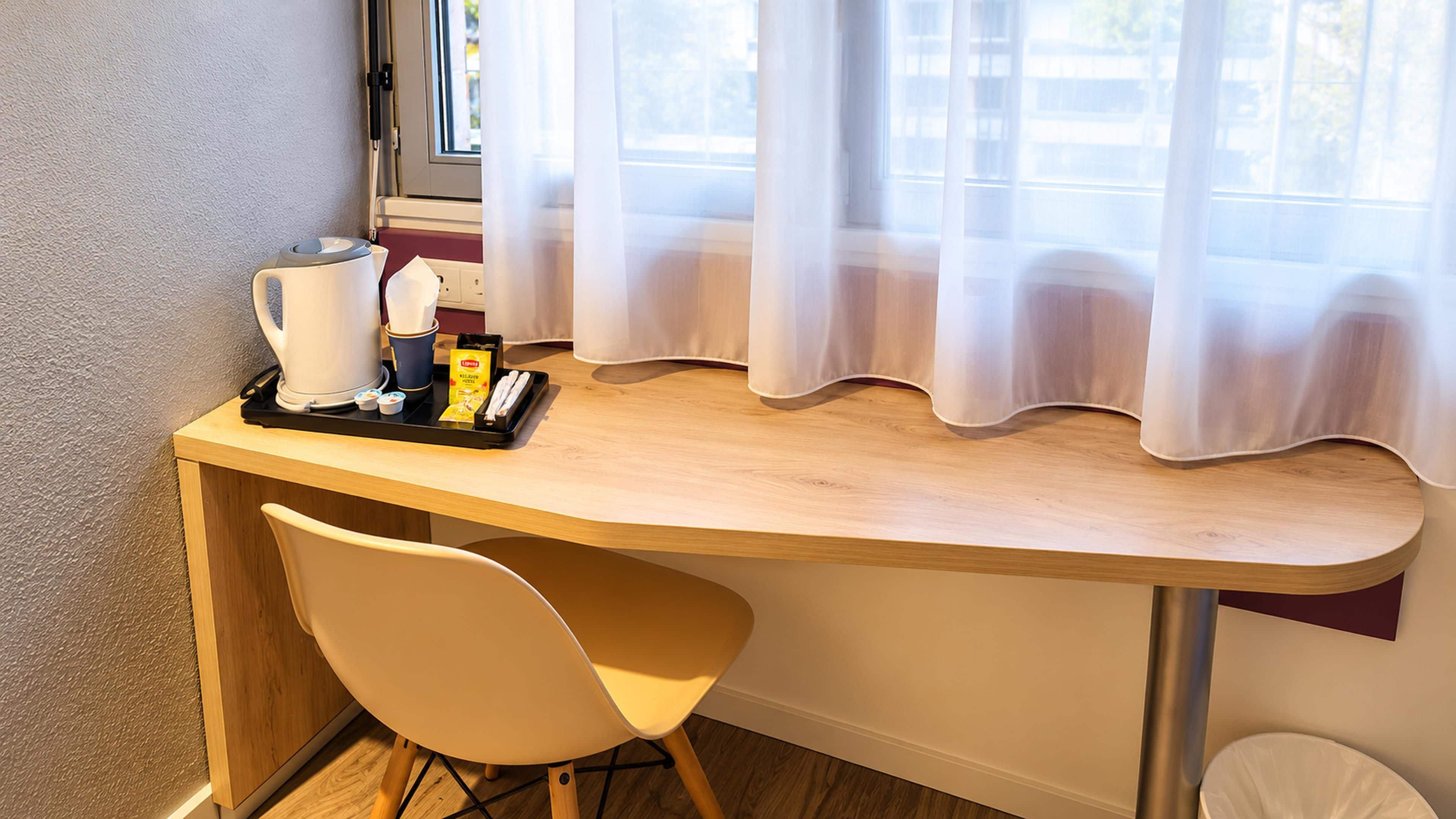 Ein kompakter Raum mit Schreibtisch aus Holz, einem Stuhl, einem Fenster mit leichtem Vorhang und einer kleinen Mülltonne.