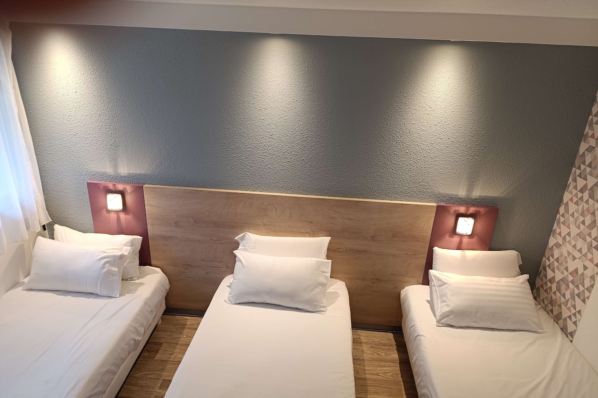 Une chambre d’hôtel avec trois lits simples, chacun doté de draps et d’oreillers blancs. Les têtes de lit sont en bois et comportent des lampes intégrées fixées au mur. La chambre possède un mur gris texturé et un plancher en bois.
