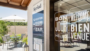 Entrada de hotel com balcão de recepção, área de estar externa e placa indicando um bar de café da manhã.
