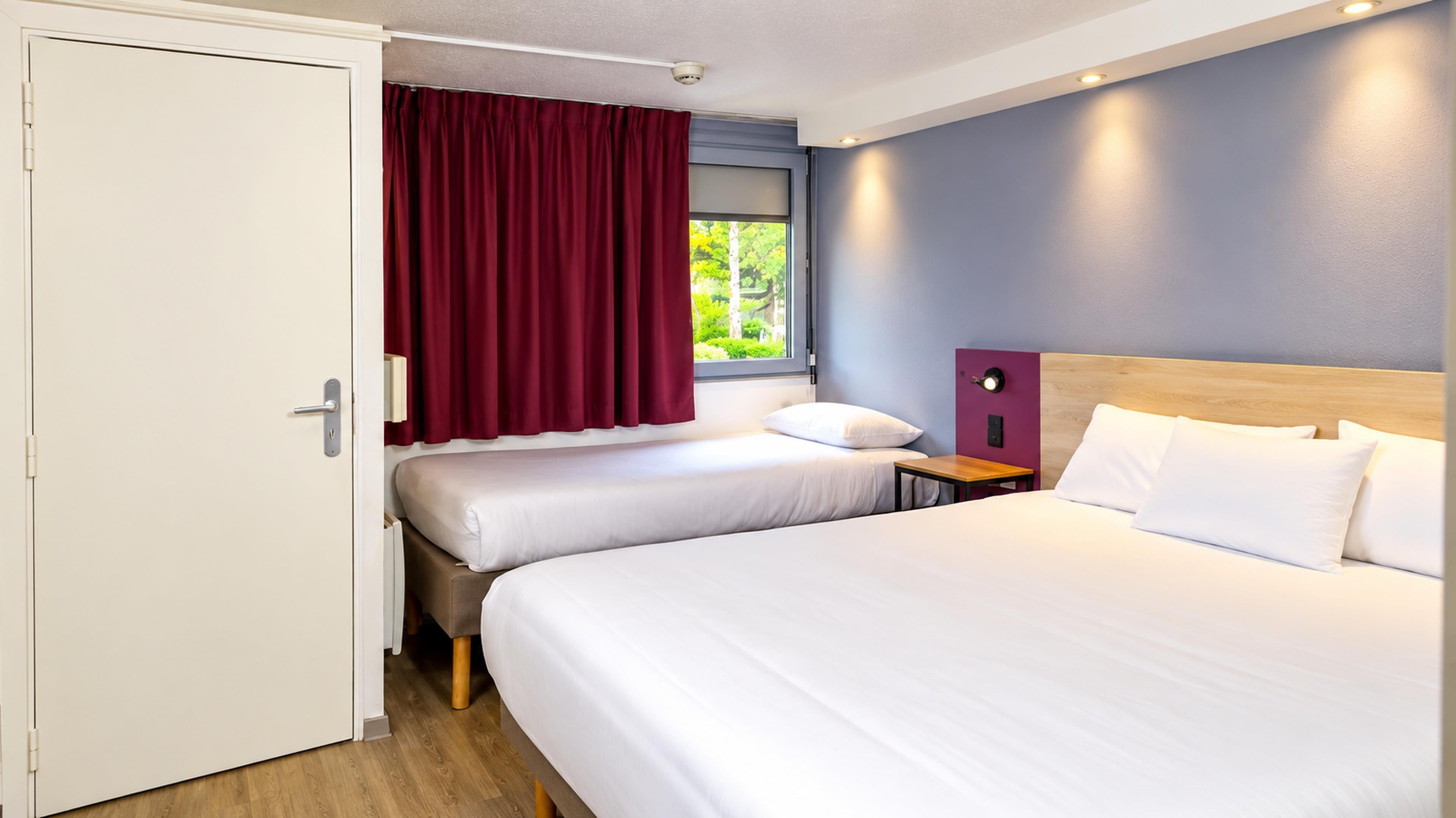 Une chambre d’hôtel avec deux lits, une fenêtre avec des rideaux, une table de chevet et un luminaire fixé au mur.